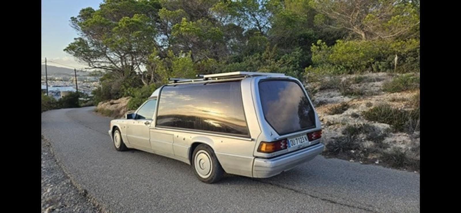 Silver Mercedes hearse conversion with solar on roof rack, parked on a winding Spanish coastal road overlooking a harbor